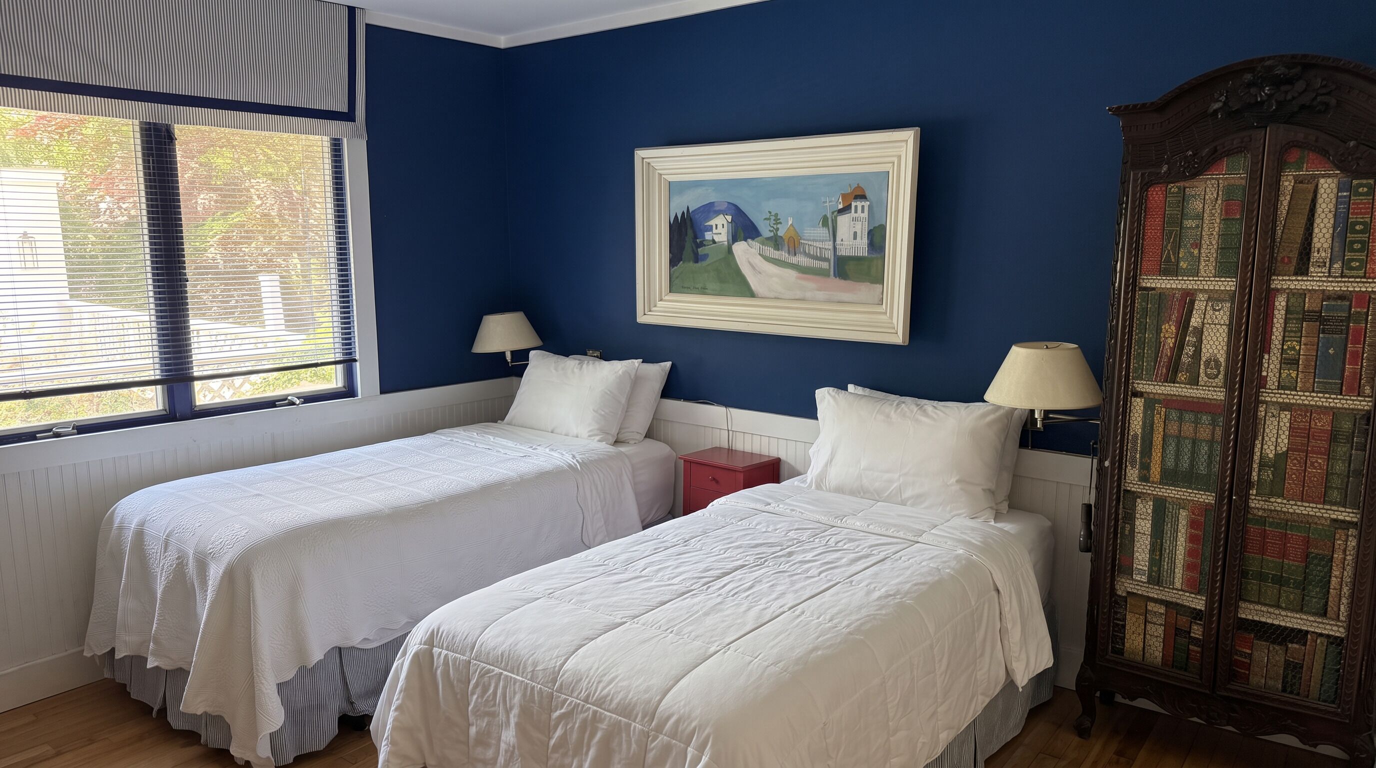 Um quarto com duas camas de solteiro, uma janela com persianas, um quadro na parede e uma estante de livros.