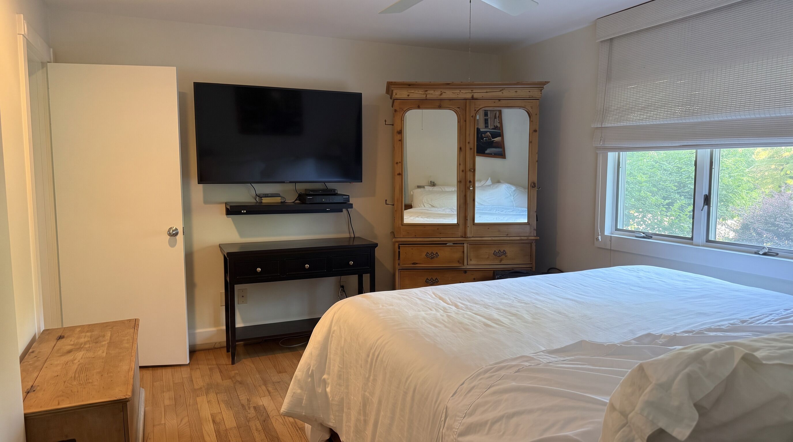Een slaapkamer met een bed, een aan de muur bevestigde televisie, een houten dressoir en een raam met zonwering.