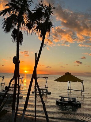 Eine Strandlandschaft mit Palmen, einem Sonnenuntergang und einem kleinen Steg mit Tisch und Stühlen.