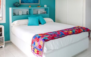 A bed with white bedding and a colorful quilt, a teal wall, and a shelf with various items.