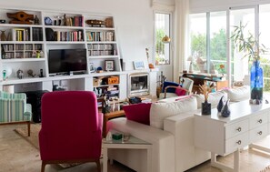 A living room with a white sofa, a red chair, a bookshelf, a television, and a window with a view of trees. - Gorgeous home in Mojacar with private swimming pool, can be inside or outside (Mojacar)