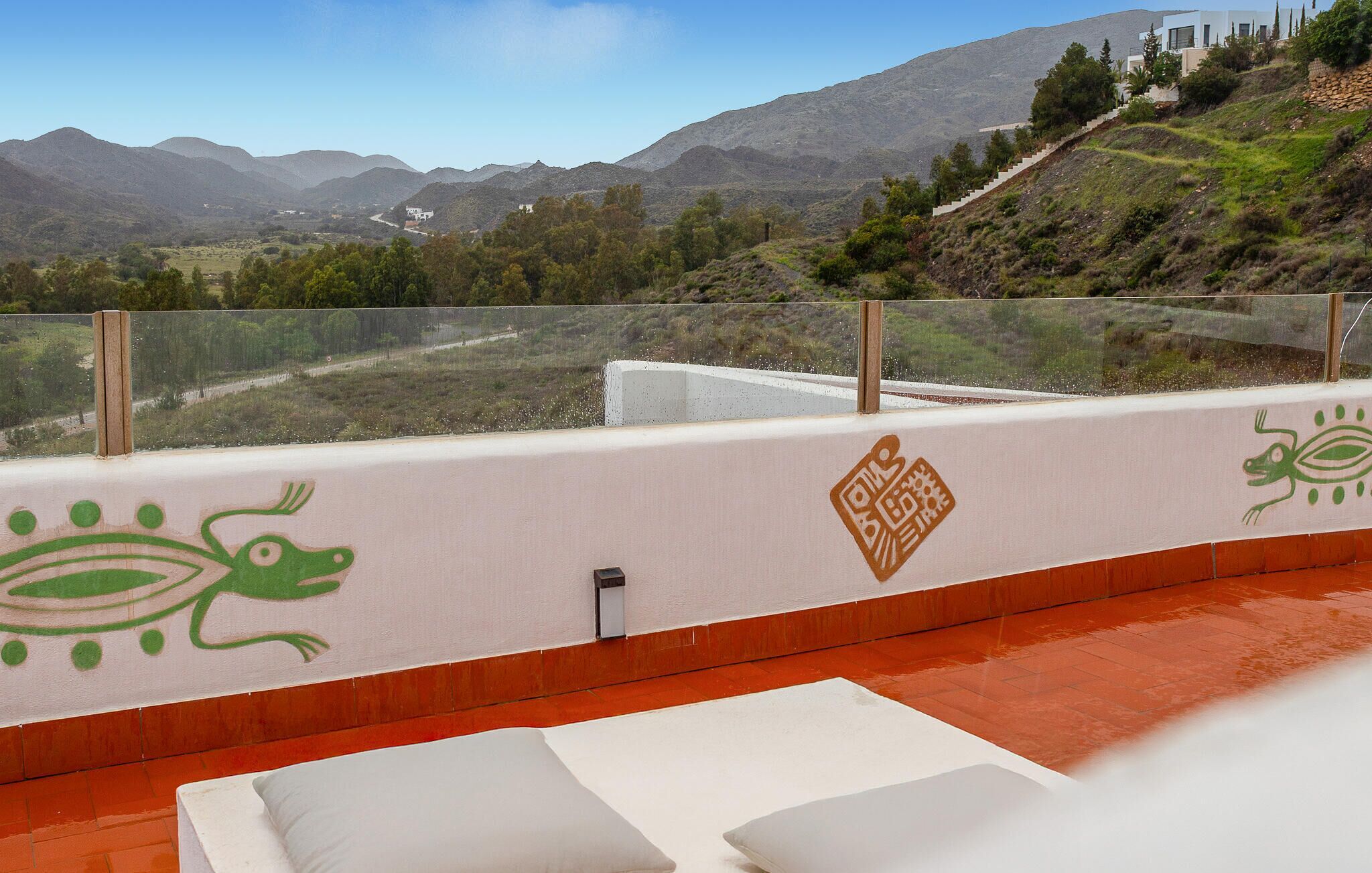 Un balcón con vistas a montañas, una pared blanca con motivos decorativos en tonos verde y marrón, y un suelo de baldosas rojas.