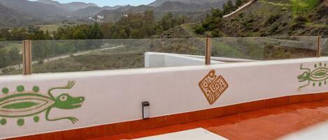 Un balcón con vistas a montañas, una pared blanca con motivos decorativos en tonos verde y marrón, y un suelo de baldosas rojas.