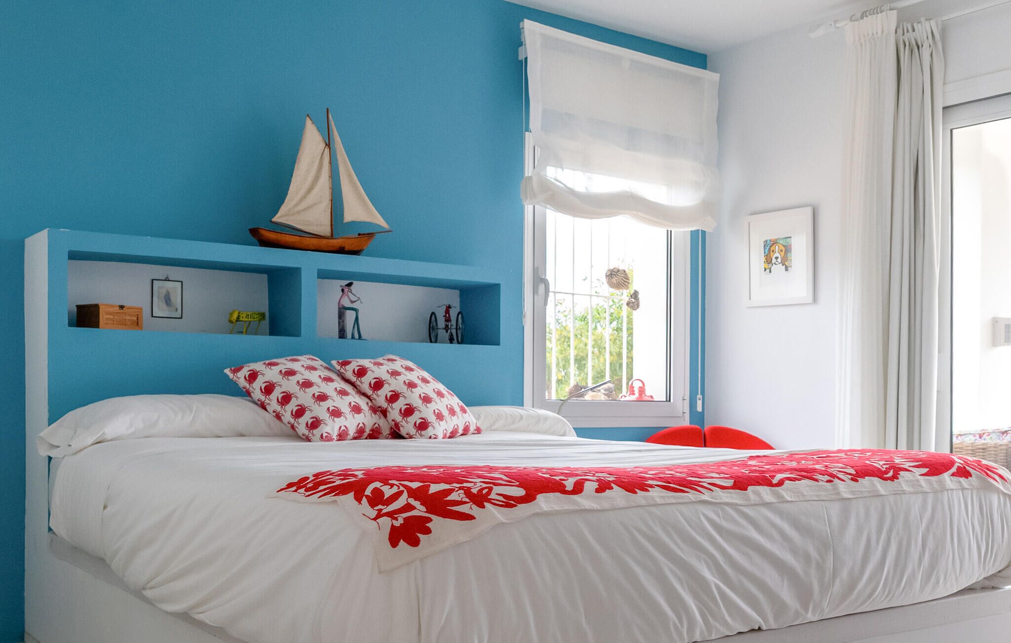 A bedroom with a bed, a sailboat decoration, a window with curtains, and a wall with shelves.