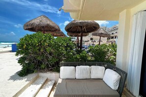 Ein Balkon mit gepolsterten Sitzgelegenheiten, umgeben von reetgedeckten Sonnenschirmen und Blick auf einen Strand mit blauem Himmel.