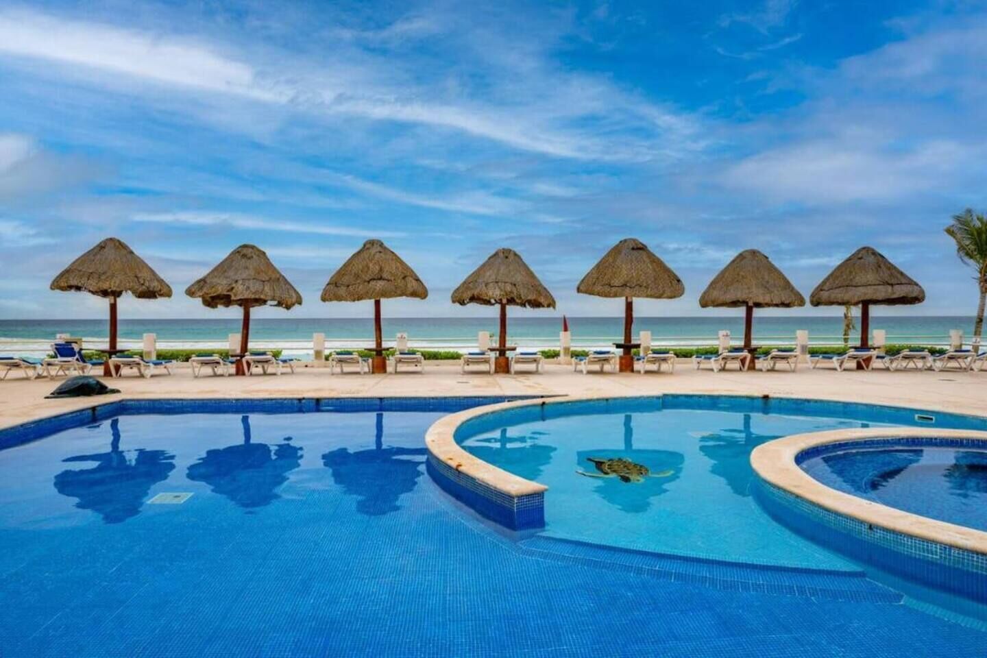 Uma piscina com borda circular, rodeada por espreguiçadeiras e guarda-sóis de palha, com vista para o oceano.
