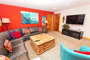A living room with a sectional sofa, a coffee table, a television, and a painting on the wall. - Oceanside, 2 Hot Tubs- 2 Duplex's rents as 1-BU22, Love R Shack at the Beach (Corolla)