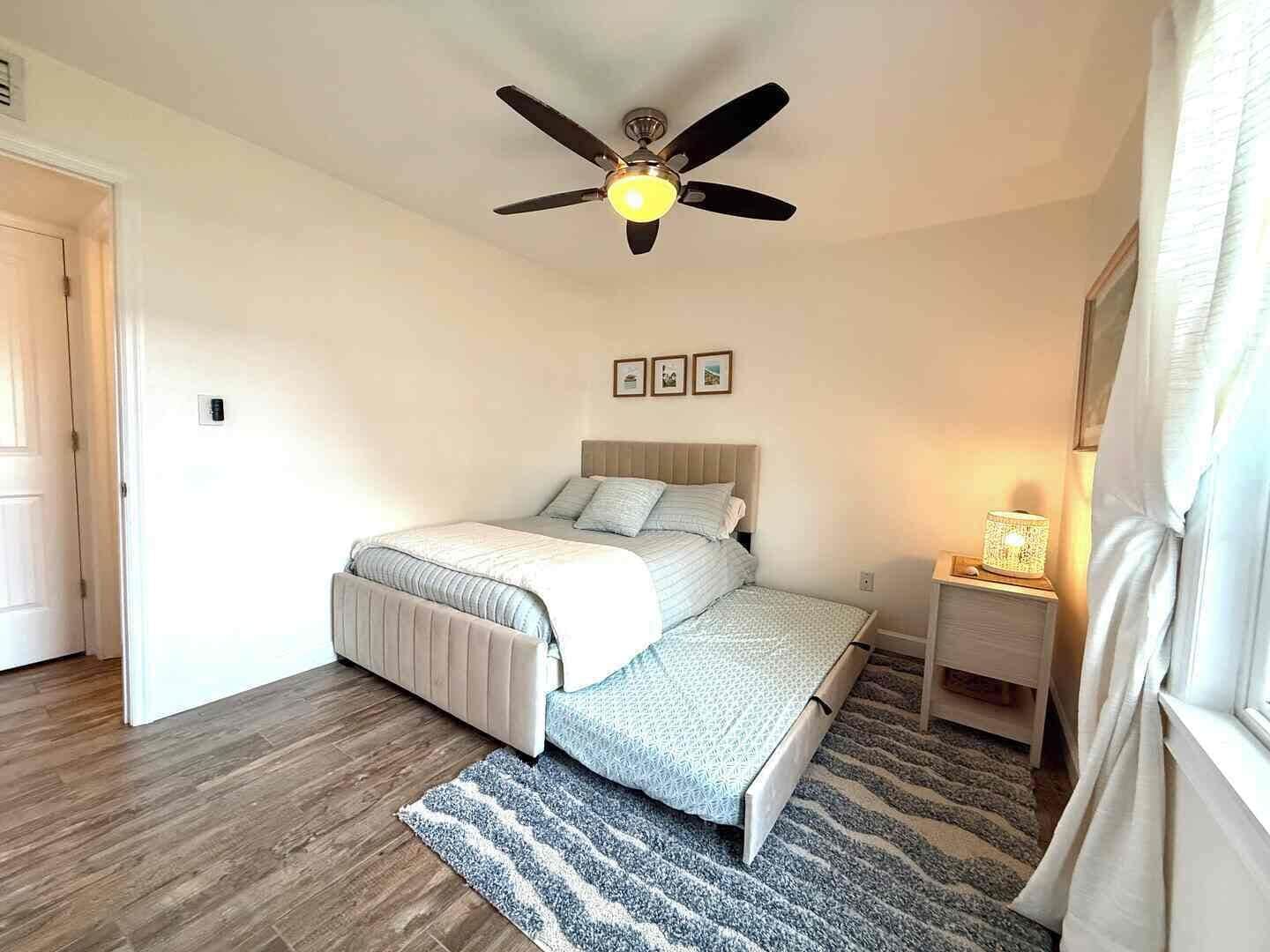 A bedroom with a bed, a ceiling fan, a nightstand with a lamp, and a window with curtains.