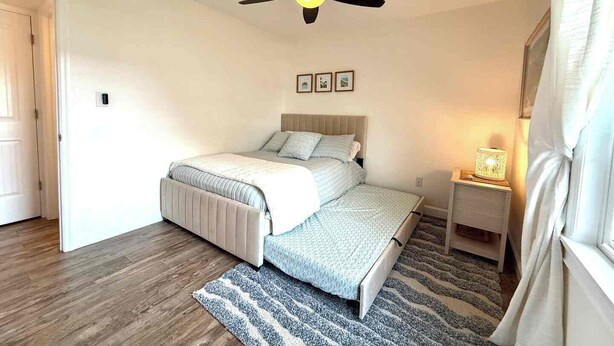 Een slaapkamer met een bed, een plafondventilator, een nachtkastje met een lamp en een raam met gordijnen.