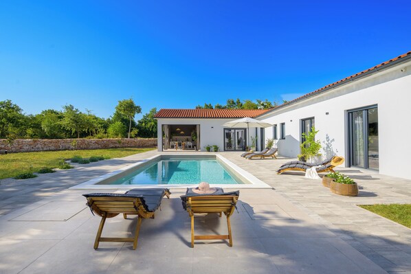 Une maison moderne dotée d’une piscine, de deux fauteuils de jardin en bois et d’un espace patio.