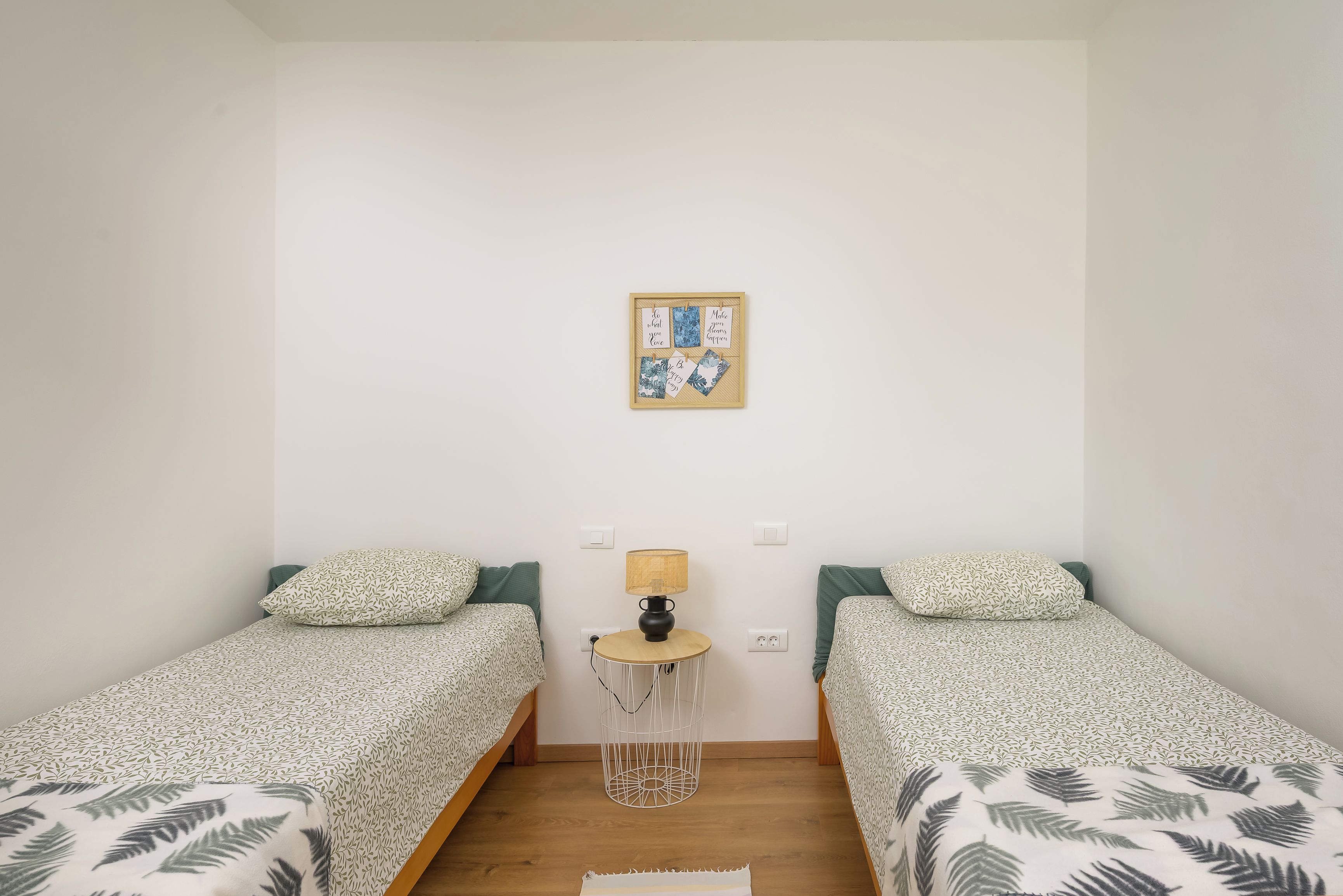 A room with two single beds, a small table with a lamp, and a wall-mounted picture frame with inspirational quotes.
