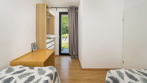 Una habitación pequeña con cama, un banco de madera, un armario y vistas por una puerta.
