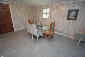 A dining area with a glass-top table surrounded by wicker chairs, a wooden door, and a window with blinds. - Kismet - Oceanfront in Garden City Beach (Garden City Beach)