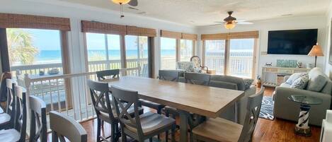 A dining area with a wooden table and chairs, a ceiling fan, and large windows with a view of the ocean.