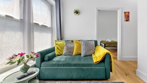 Ett modernt vardagsrum med en grön soffa, ett litet sidobord med en vas och en fjärrkontroll, och utsikt över ett sovrum genom en öppen dörr.