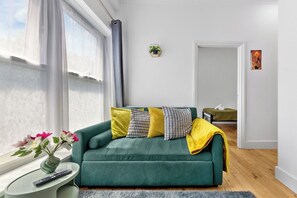 A modern living room with a green sofa, a small side table with a vase and remote control, and a view of a bedroom through an open door. - Modern 2BR Apartment | Sleeps 5 | Canary Wharf E14 (London)