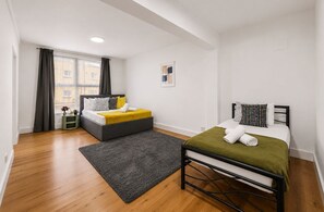 A modern bedroom with a bed, a smaller bed, a window, and a grey rug. - Modern 2BR Apartment | Sleeps 5 | Canary Wharf E14 (London)