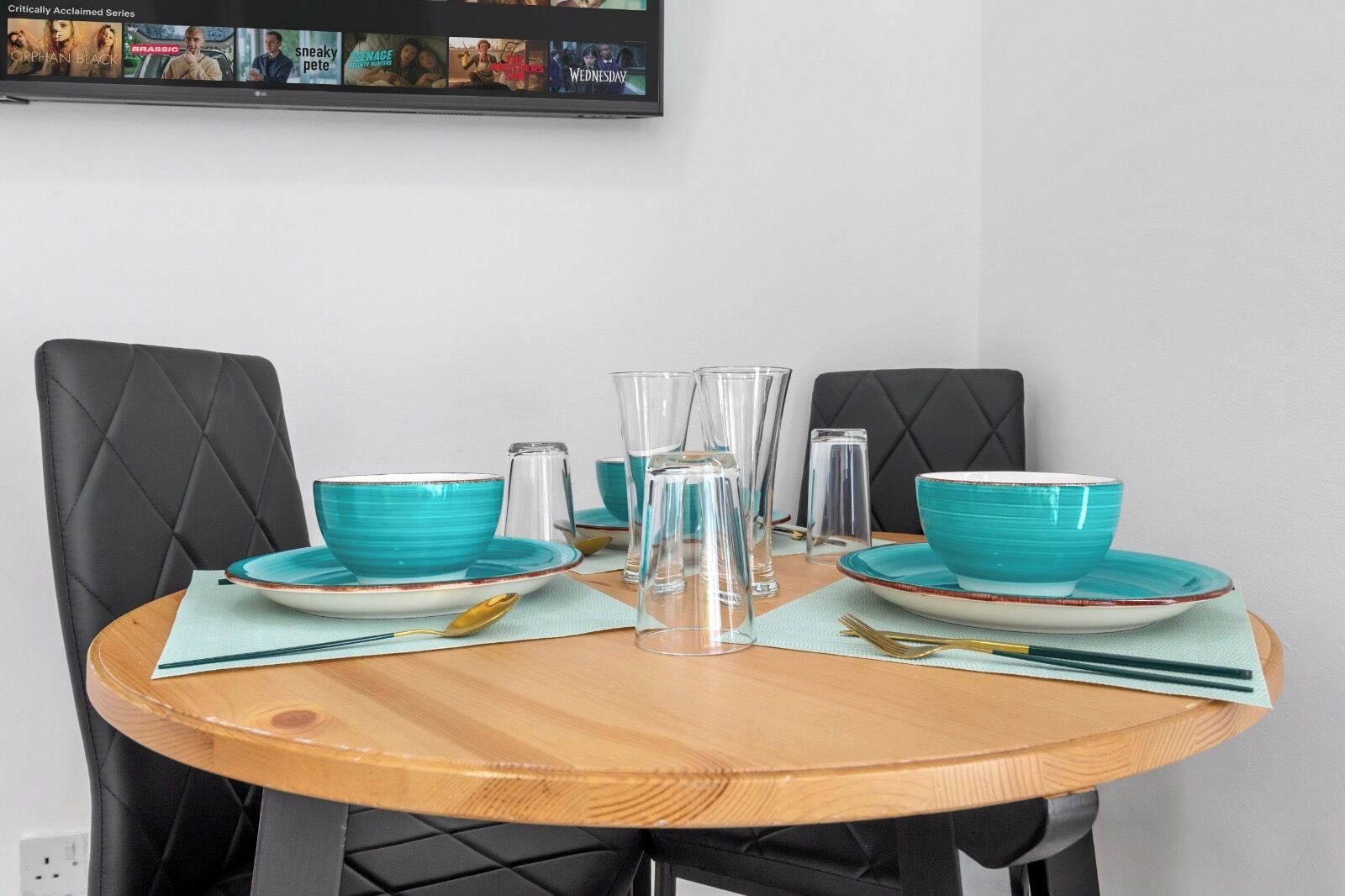 A round wooden dining table with turquoise bowls and clear glasses set for two.