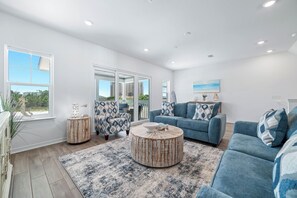 A spacious living room with a large window, a blue sofa set, a round coffee table, and a patterned rug. - Luxury Lost Key Retreat | Private Beach Club, Pools & Golf | BEACHBALL PROPERTIES (Pensacola)