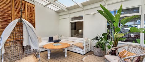 Un patio soleado con una terraza de madera, una silla colgante, un banco acolchado y una silla de mimbre.