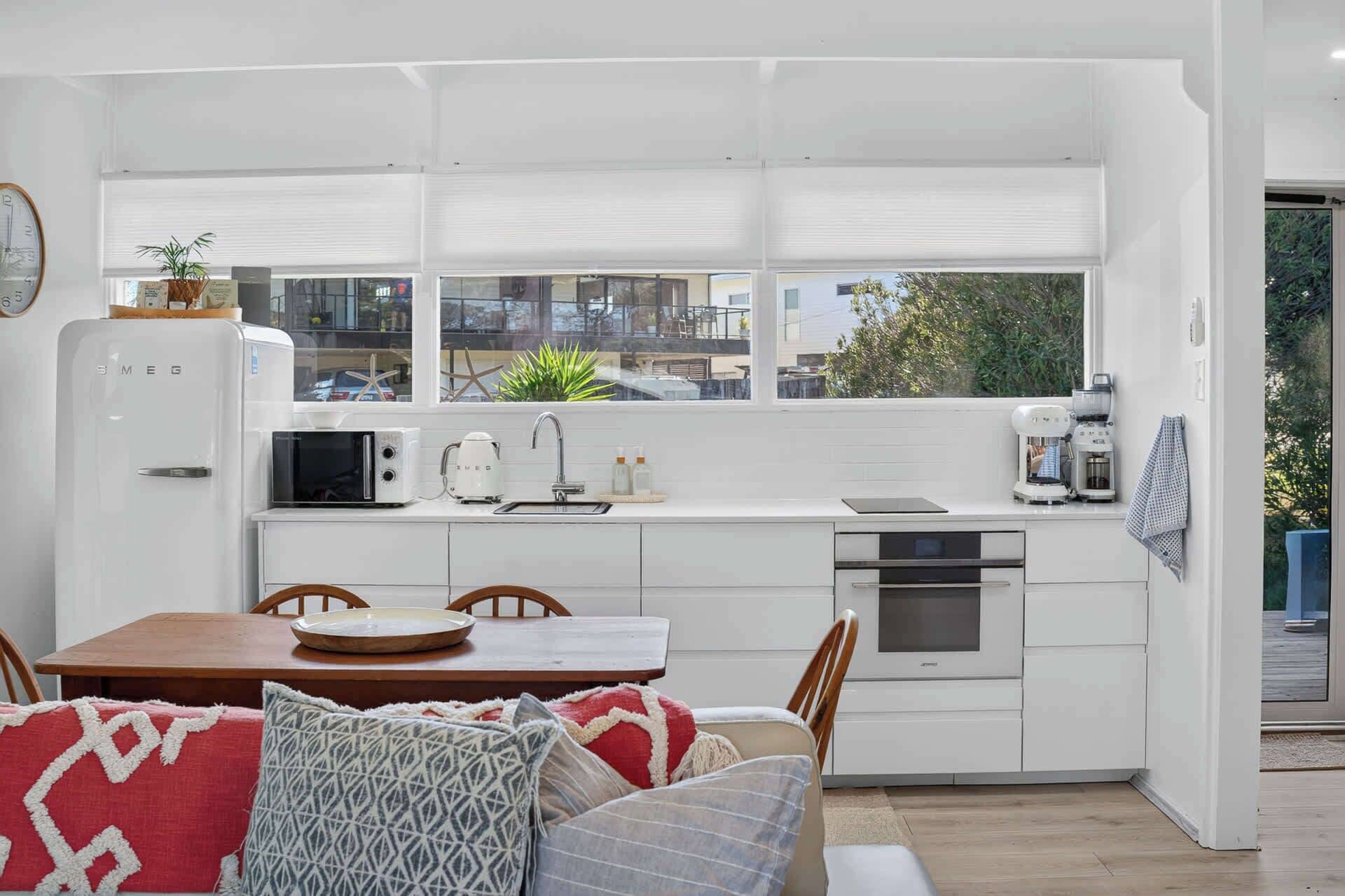 A compact kitchen with white cabinetry, a microwave, a kettle, and a coffee machine. There is a dining area with a wooden table and chairs, and a window with blinds.