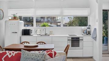 A compact kitchen with white cabinetry, a microwave, a kettle, and a coffee machine. There is a dining area with a wooden table and chairs, and a window with blinds.