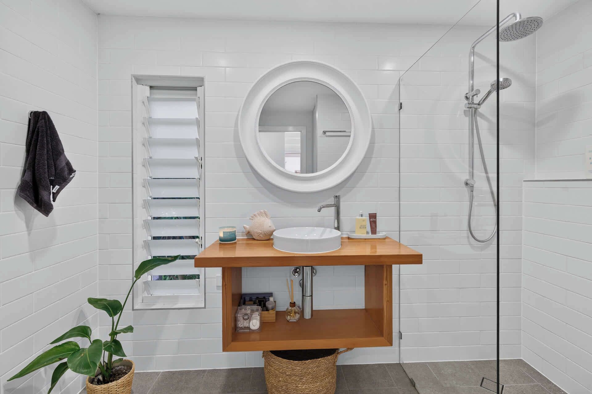 A modern bathroom with a white tiled wall, a round mirror, a wooden vanity, a sink, a shower, and a towel rack.