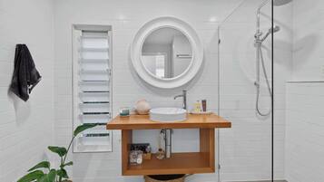 A modern bathroom with a white tiled wall, a round mirror, a wooden vanity, a sink, a shower, and a towel rack.