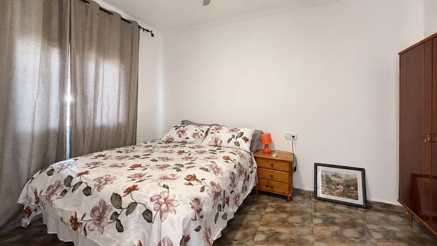 Ein Schlafzimmer mit einem Bett, einem Nachttisch, einer Lampe, einem gerahmten Bild, einem Holzschrank und einem Deckenventilator.