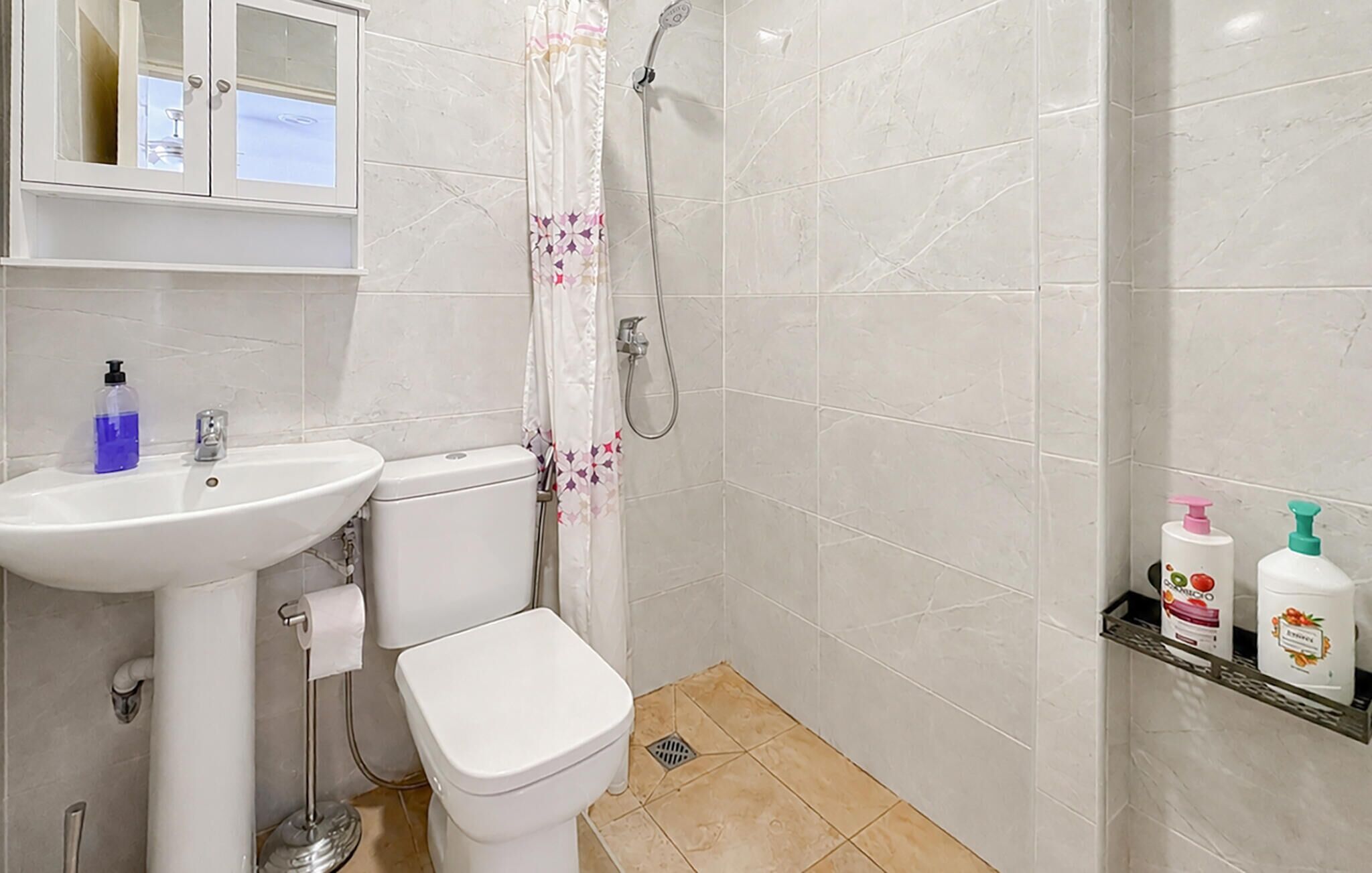 A bathroom with a toilet, sink, shower, and a shelf with toiletry bottles.