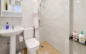 Une salle de bain équipée de toilettes, d’un lavabo, d’une douche et d’une étagère où sont rangées des bouteilles de produits de toilette.