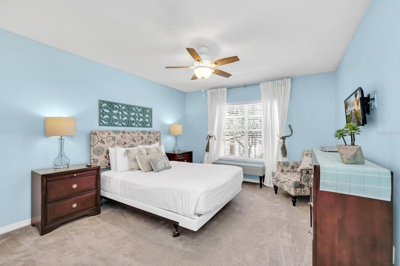 A bedroom with a bed, nightstands, a desk, a ceiling fan, and a window with curtains.