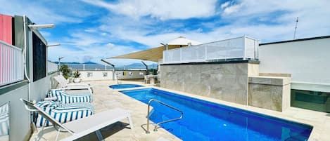 A swimming pool with lounge chairs and a sunshade, surrounded by a tiled deck and a building with a white facade.
