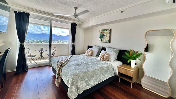 Un dormitorio con cama, mesita de noche, planta en maceta, ventilador de techo y vistas al exterior.