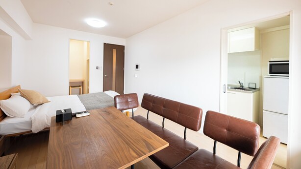 Une chambre d’hôtel avec une table à manger en bois, deux fauteuils en cuir marron, un lit avec une literie blanche, une petite table de chevet en bois et une kitchenette équipée d’un four à micro-ondes.