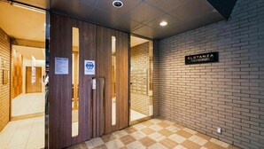 L’entrée d’un hôtel, avec une porte en bois, une enseigne indiquant « ELSTANZA TENJINHIGASHI » et un comptoir d’enregistrement visible à travers les portes vitrées.