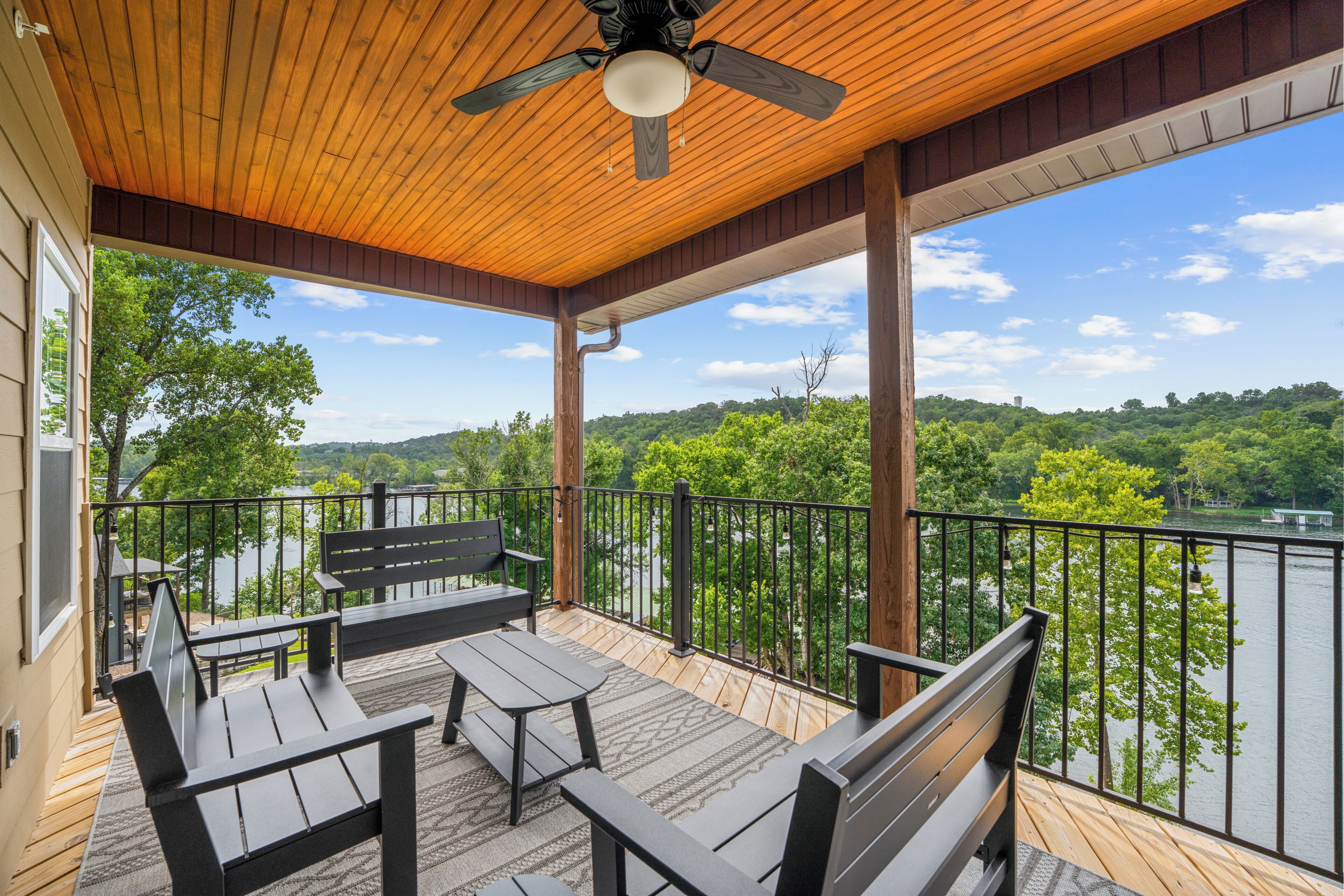 A covered outdoor deck with wooden flooring, a ceiling fan, and a view of a lake surrounded by trees.