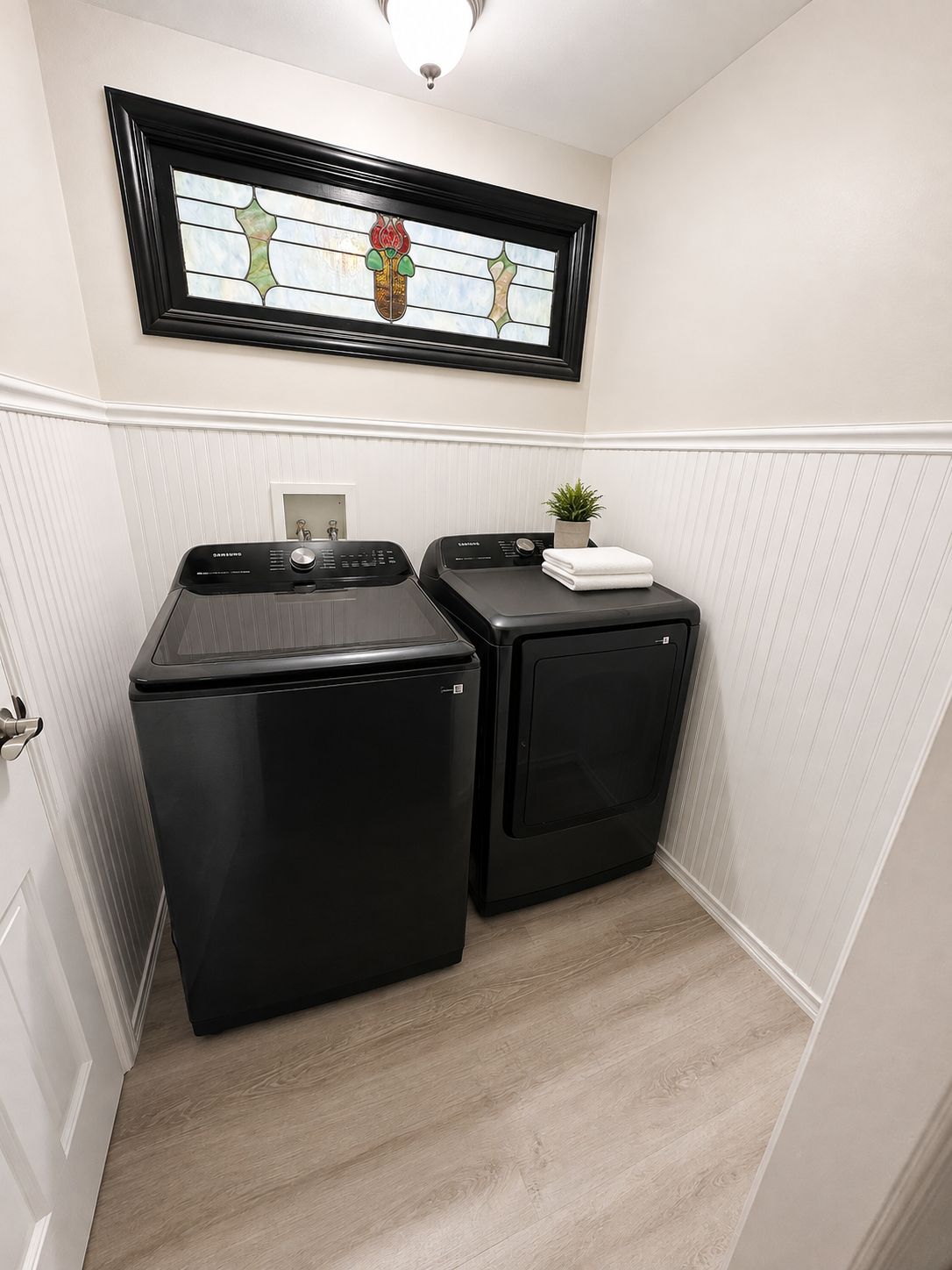 A laundry room with two black appliances, a framed stained glass window above, and a small plant on the right.