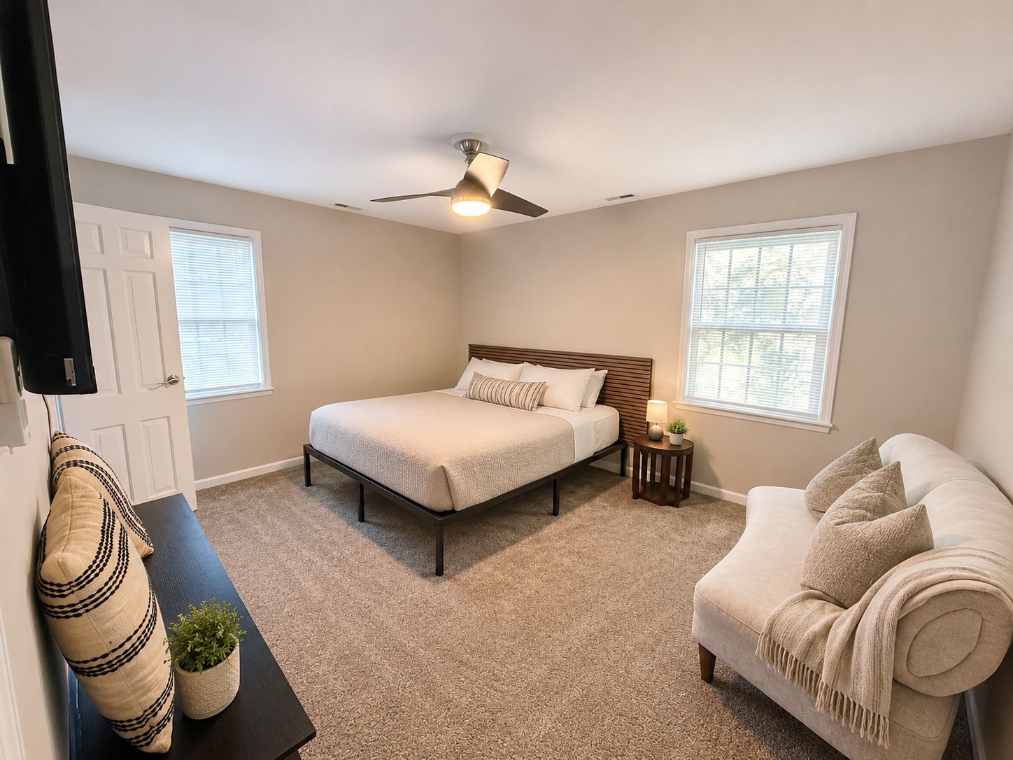 A bedroom with a bed, a ceiling fan, a bedside table with a lamp, a window, and a couch.