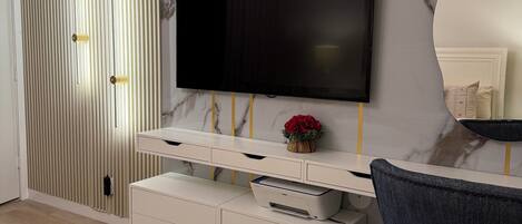 Una sala de estar moderna con un televisor de pantalla plana montado en una pared de mármol, un mueble blanco con cajones, un sillón azul y una manta color beige sobre piso de madera.