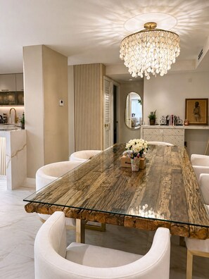 Ein moderner Essbereich mit einem Glastisch, weiß gepolsterten Stühlen, einer dekorativen Chandelierleuchte und einem Spiegel an der Wand.
