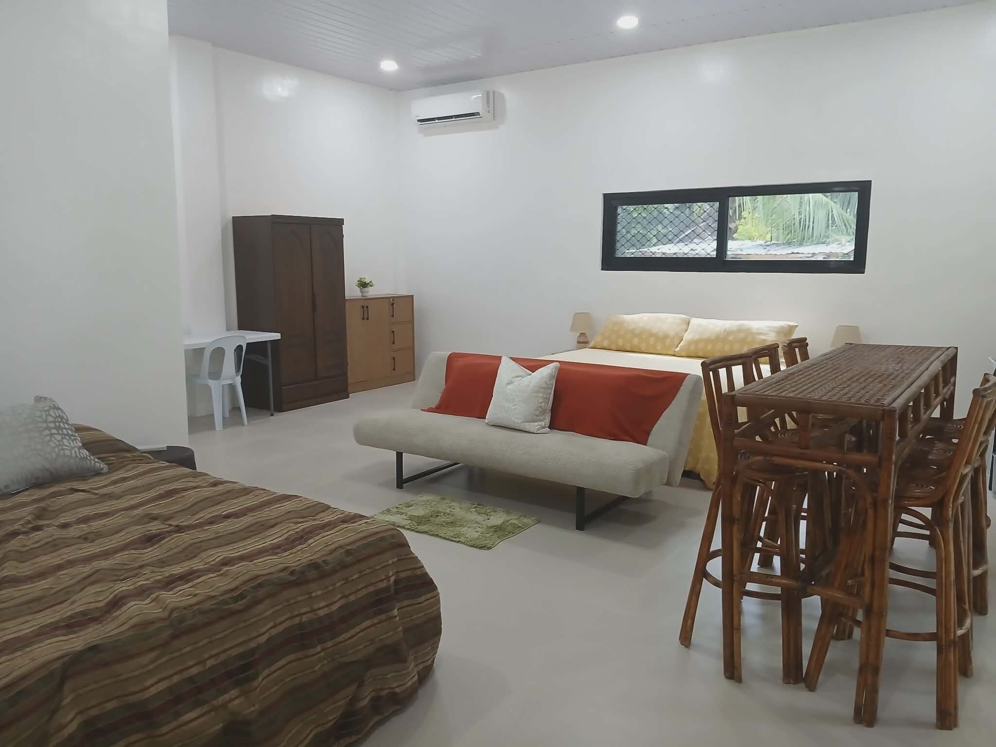 A room with a bed, a sofa, a dining table with chairs, a wardrobe, and a window.