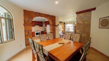Un comedor con una mesa y sillas de madera, una cocina al fondo y una pared de piedra.