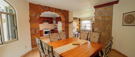 Un comedor con una mesa y sillas de madera, una cocina al fondo y una pared de piedra.