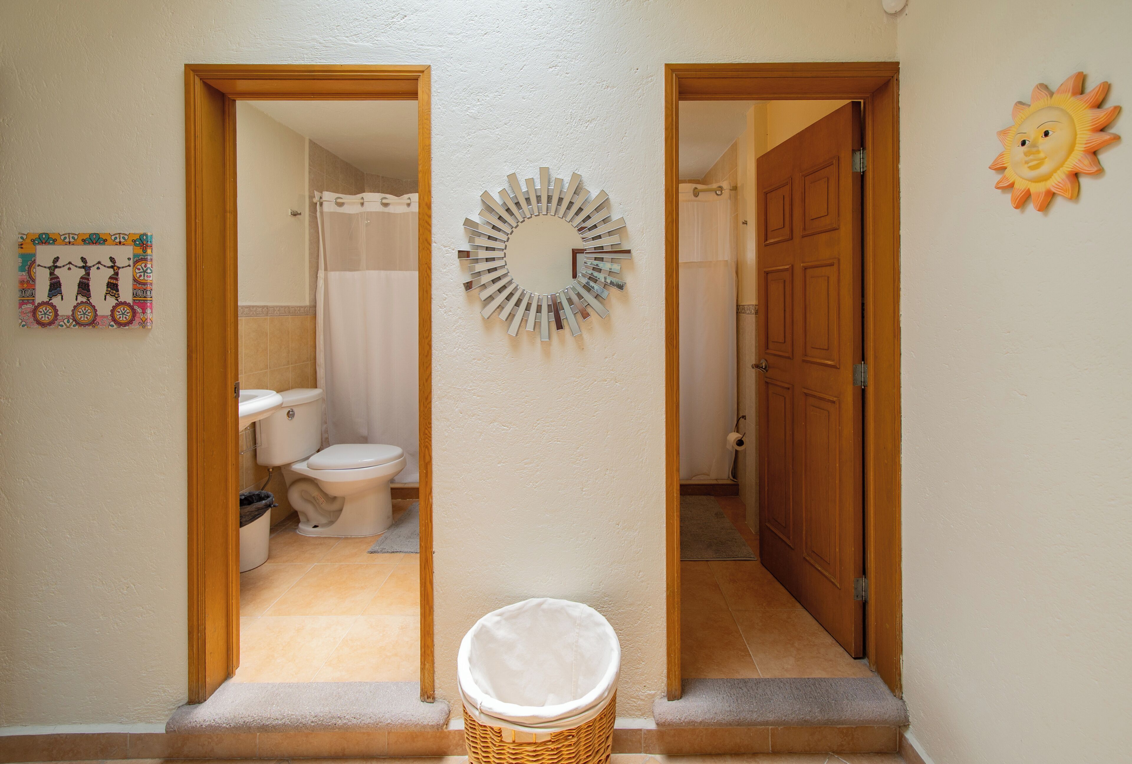 A hallway with a bathroom on the left, featuring a toilet, sink, and shower curtain. A decorative mirror hangs on the wall, and a sun decoration is visible on the right.