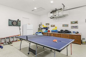A room with a ping pong table, a dartboard, a refrigerator, and a wooden cabinet with various items. - Relax, Paddle & Unwind in Cape Coral Paradise - Villa Cape Pearl - Roelens (Cape Coral)