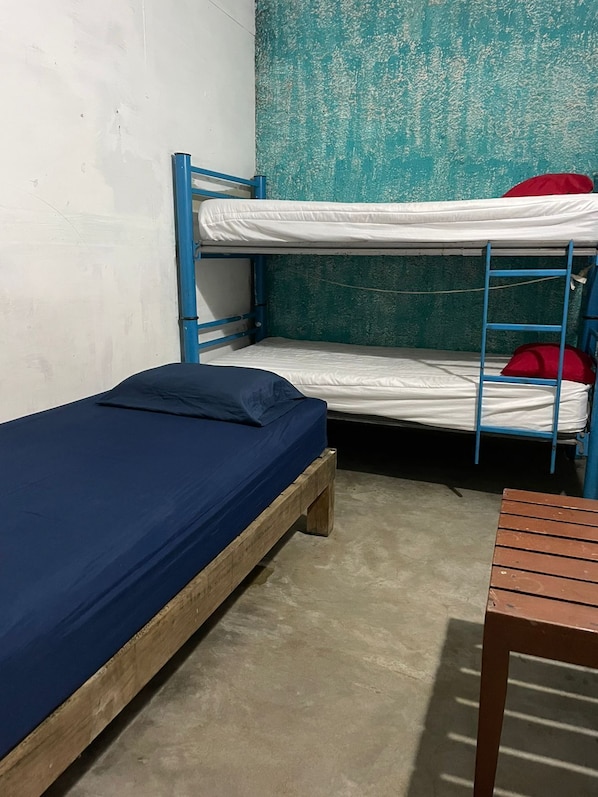 Un lit superposé avec un matelas bleu et un lit simple avec un matelas bleu, dans une pièce dont le sol est en béton et un des murs présente une texture particulière.