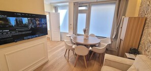 A living room with a flat-screen TV mounted on the wall, a round dining table with chairs, and a window with curtains. - Apartment - 60 sqm - Montmartre - 6p (Paris)
