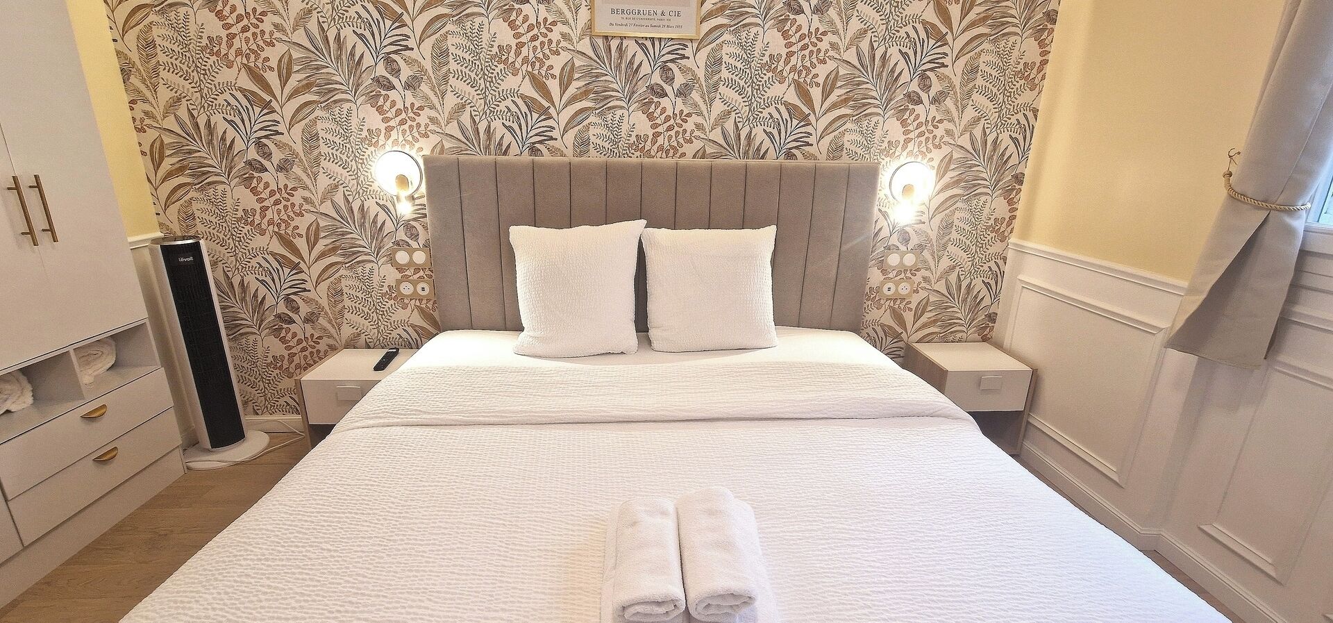 A neatly made bed with a headboard, two bedside tables with lamps, and a wall with a floral pattern.