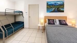 Una stanza con un letto a castello, un letto singolo, una porta, un quadro appeso al muro e un piccolo tavolo con una lampada.